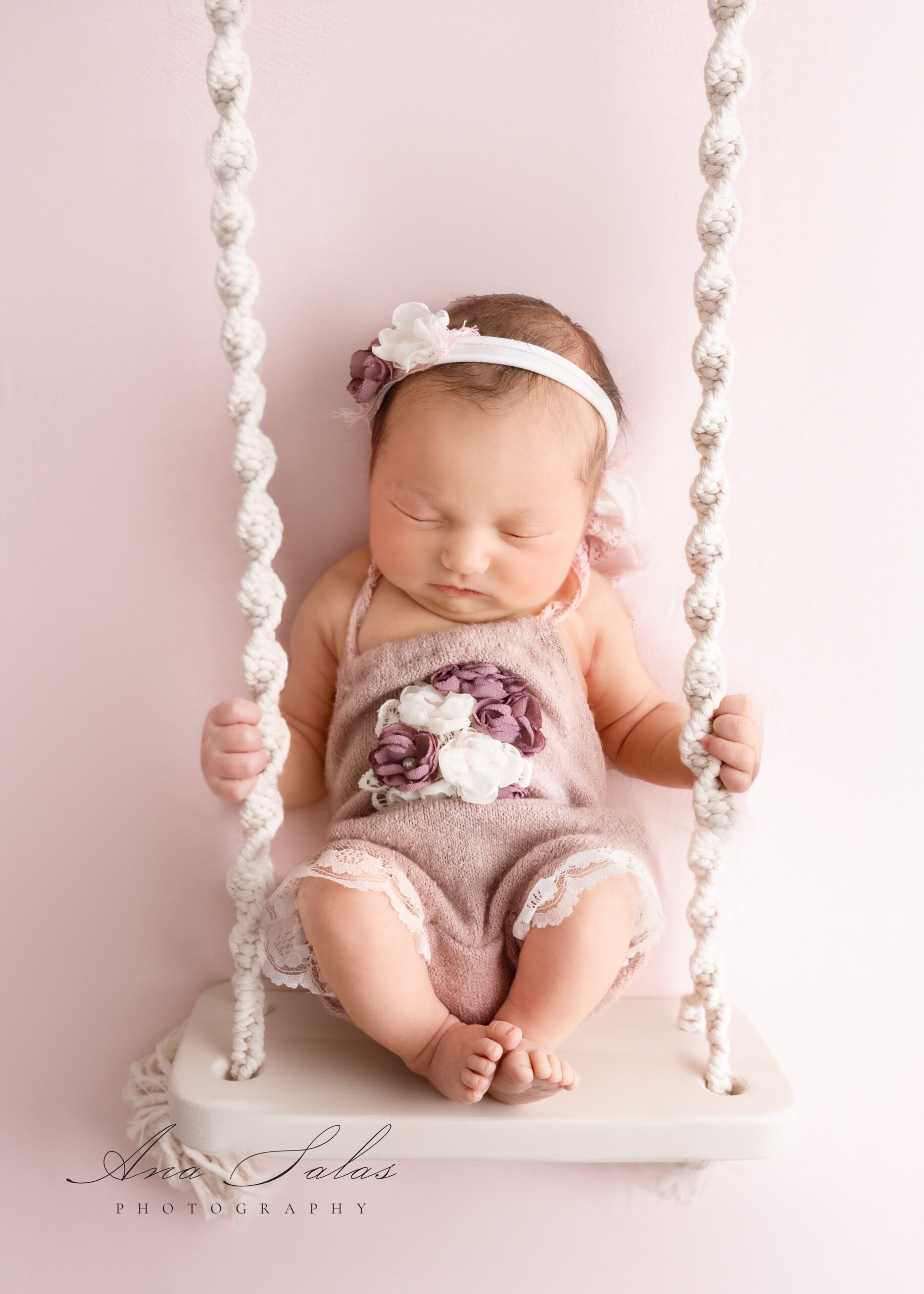 A newborn baby sleeps while sitting on a swing in a studio in a purple onesie with flowers