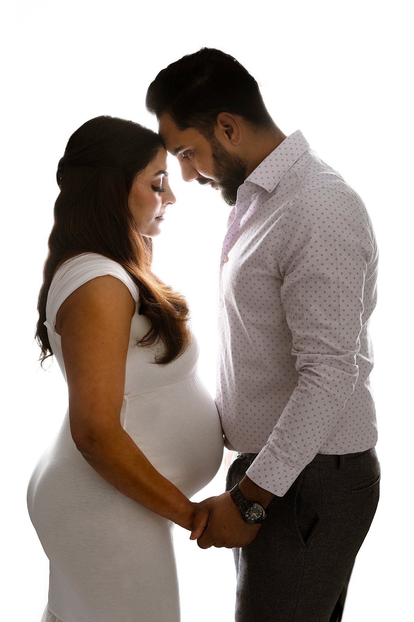 Expecting parents stand touching foreheads and holding hands in a studio in white