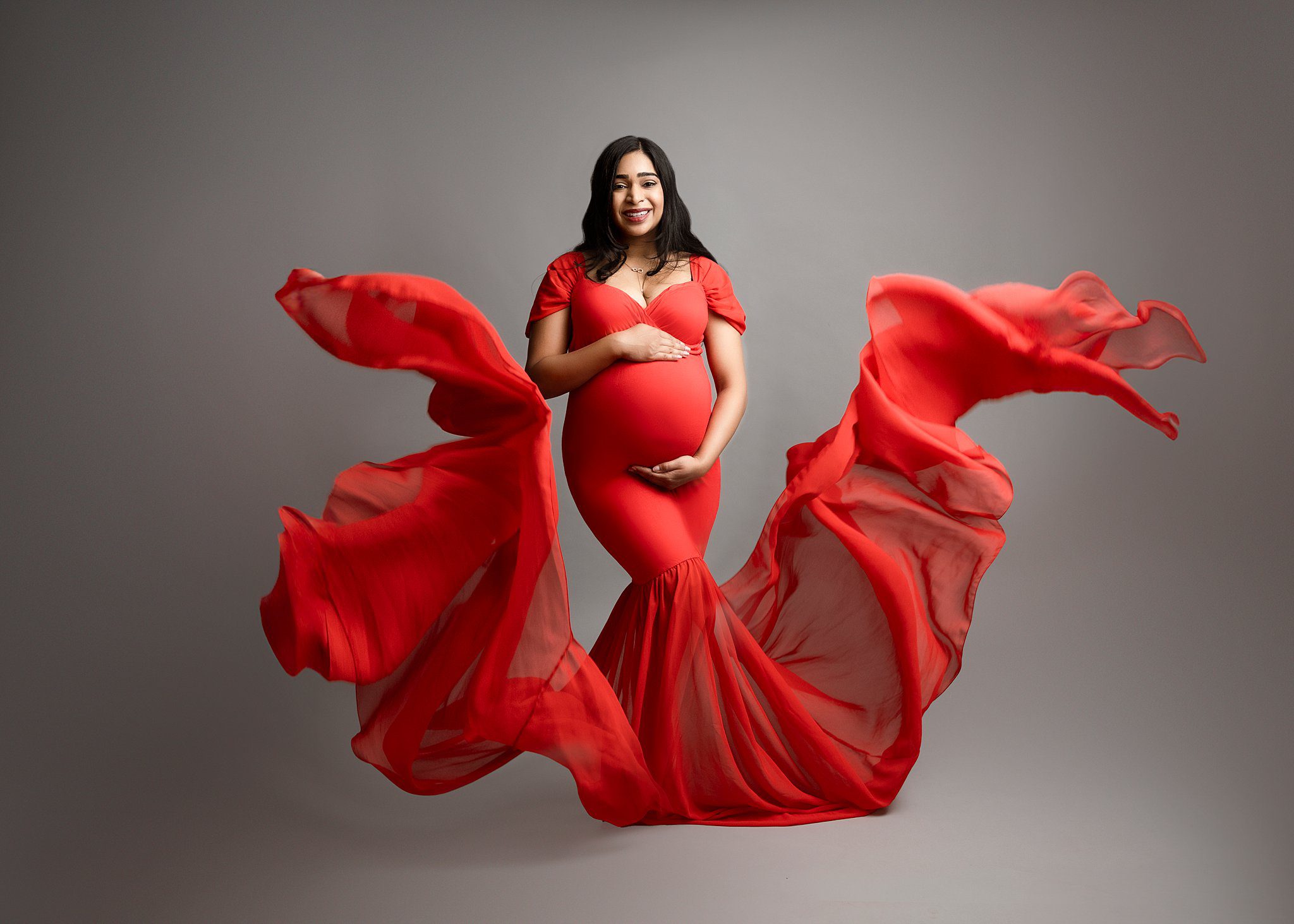 A mom to be stands in a studio in a flowing red maternity gown smiling after visiting Metta Yoga Edmonton