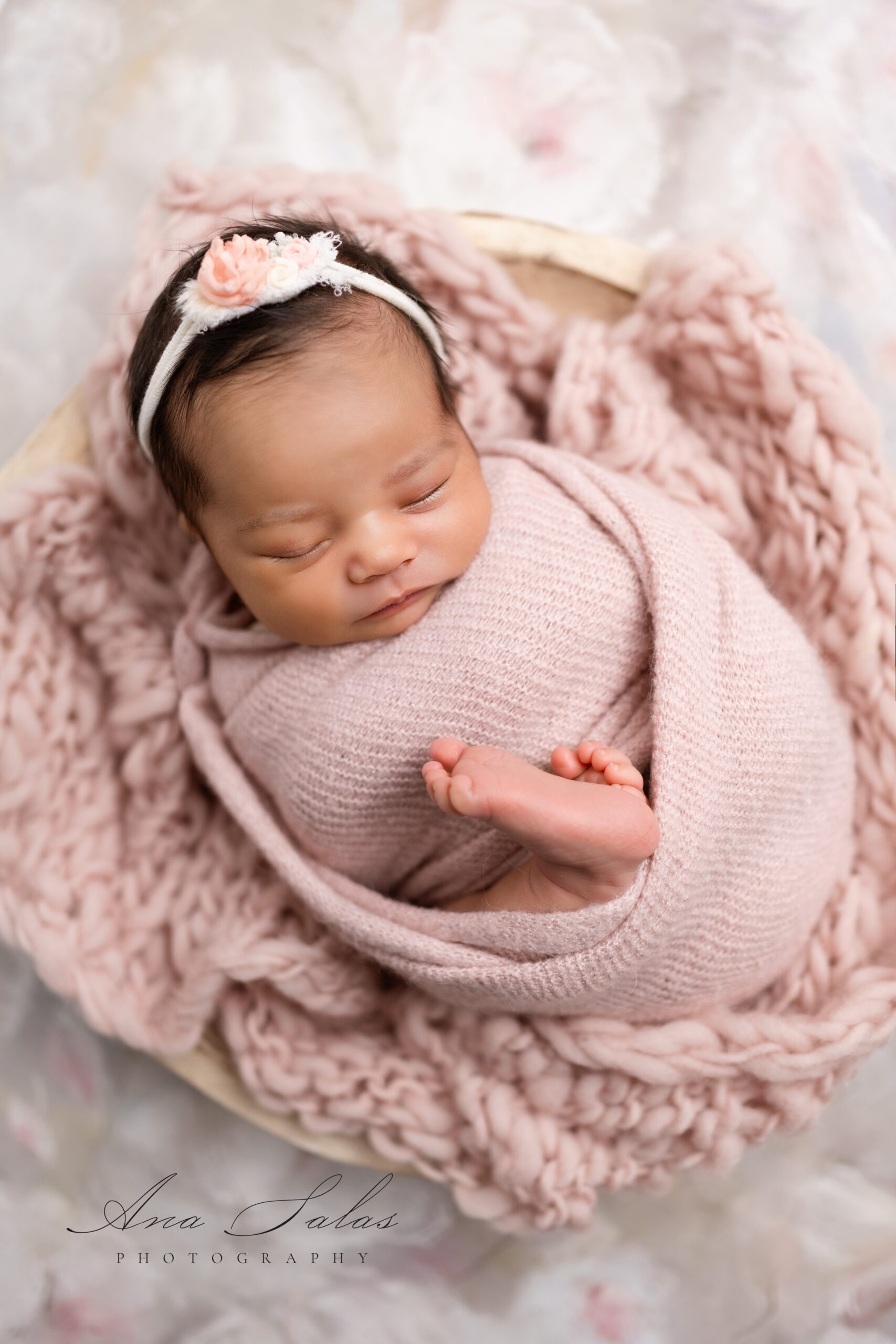 A newborn baby sleeps in a tight pink swaddle in a wooden bowl with a flower headband after meeting mini dreamers edmonton