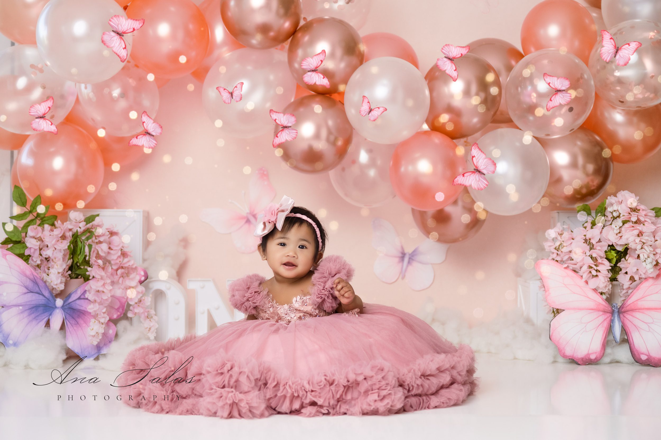 A baby girl sits in a studio in a large pink gown surrounded by butterflies