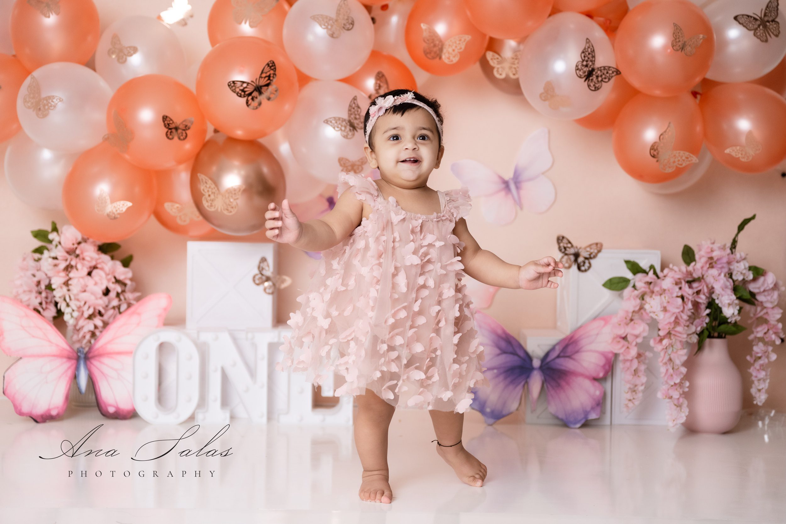 A happy baby in a pink butterfly dress smiles and stands in a studio for her birthday after visiting princess and the pea boutique