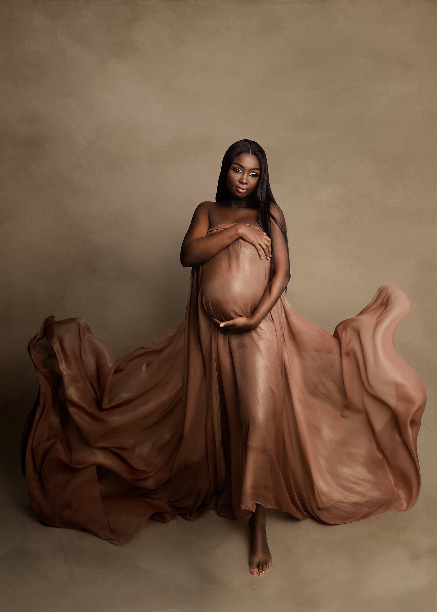 A pregnant woman walks in a studio while covering with a brown sheet as it flows around her