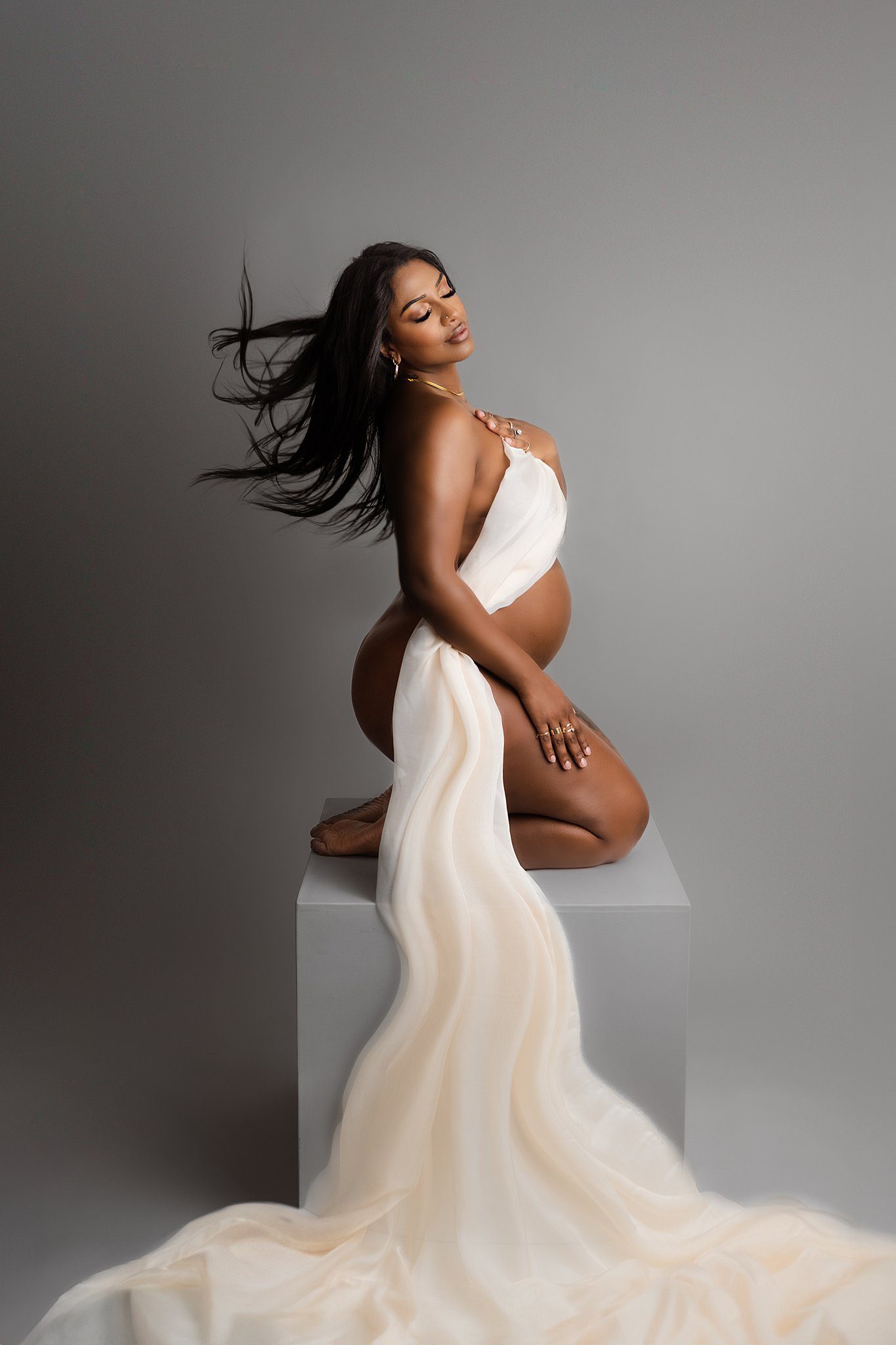 A mother to be kneels on a white box in a studio draped in a white sheet with her hair blowing behind her after visiting yoga within edmonton