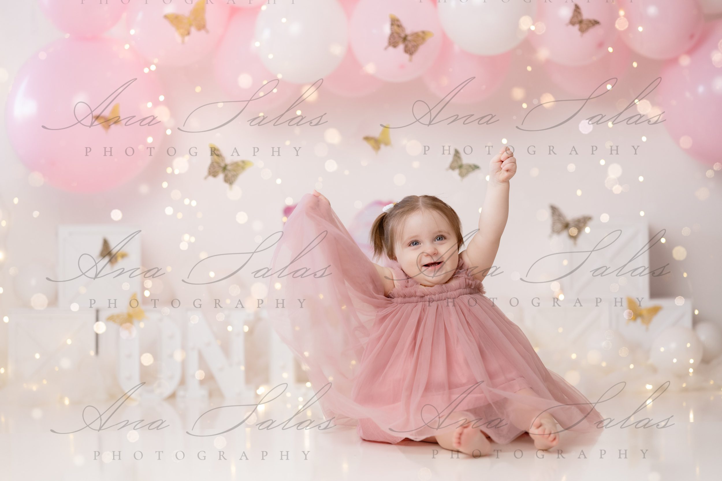 A happy baby girl in a pink dress celebrates with arms up for her birthday with butterflies after visiting head shoulders knees and toes edmonton